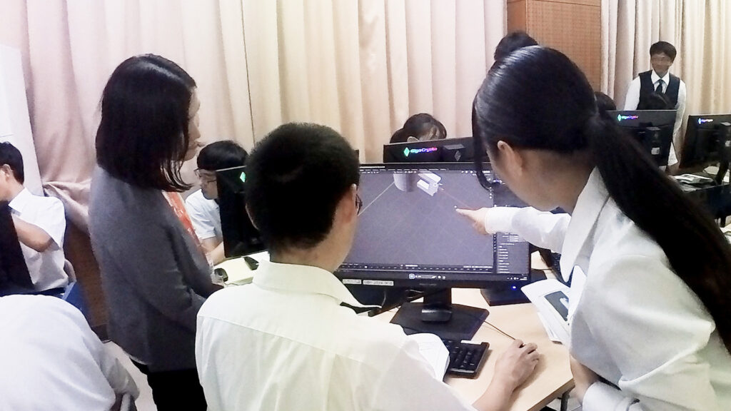 高校のコンピュータ室にて、大学生が高校生にBlenderの基礎技能をレクチャーしている様子