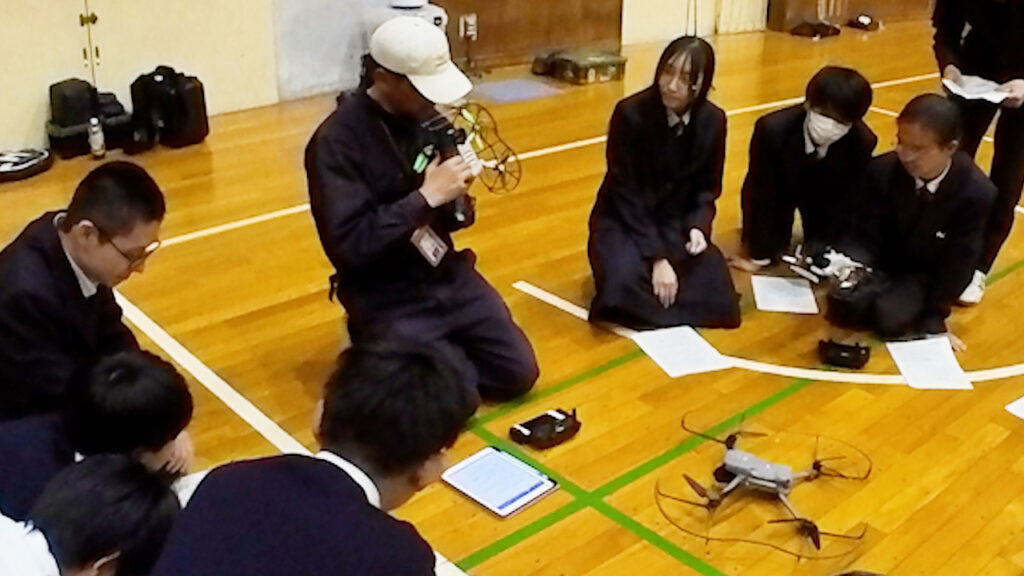 高校体育館で、講師によるドローン安全点検を高校生が観察している様子
